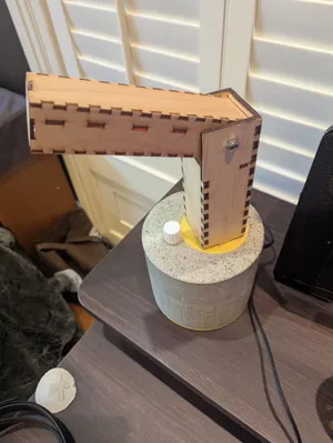 Assembled desk lamp on a nightstand: the laser-cut finger-jointed wood arm mounted on the cast-concrete cylindrical base, with the white twist-knob switch visible at its foot